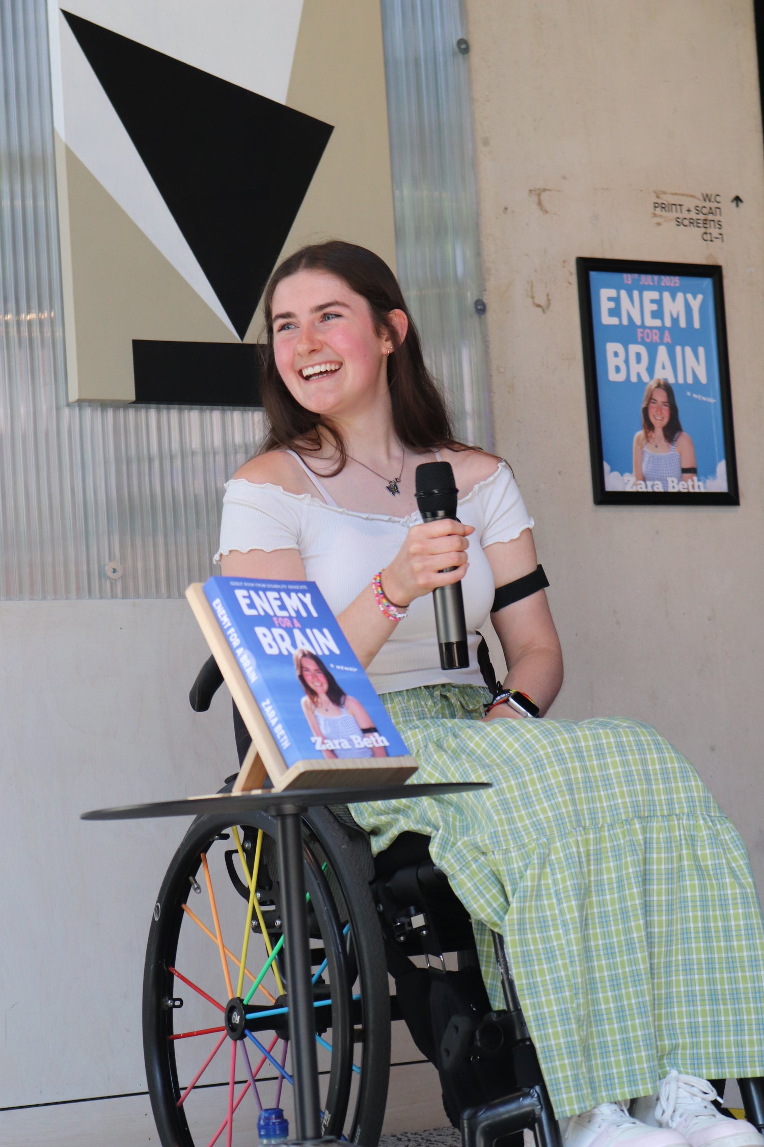 Zara Beth sat, smiling, holding a microphone next to a table with a book titled 'Enemy of the Brain'. This is a photo from her book launch event.